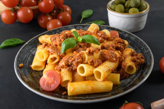 Pasta mit Bolognese-Soße, Kirschtomaten und frischem Basilikum auf schwarzem Hintergrund.