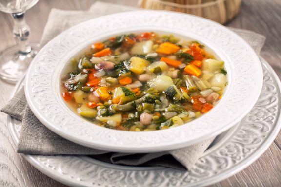 Gemüsesuppe mit buntem Gemüse in einer weißen Schüssel, serviert auf einem grauen Platz.