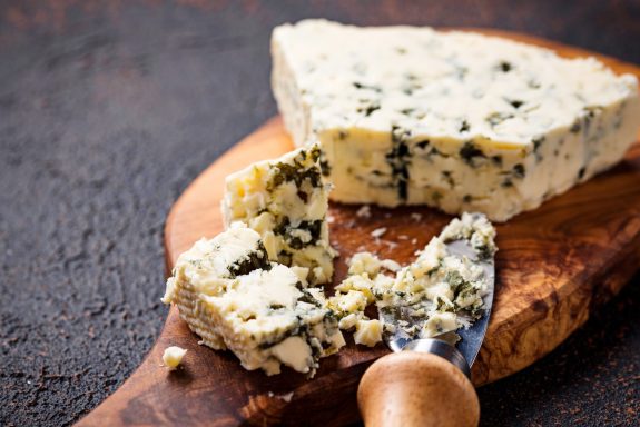 Blauschimmelkäse auf einem Holzbrett, teilweise zerbrochen, mit einem Käsemesser daneben.